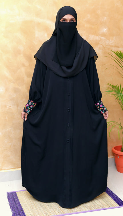 Woman wearing a black abaya with floral sleeves against a beige wall.