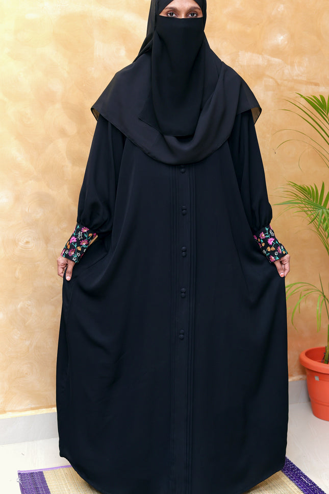 Woman wearing a black abaya with floral sleeves against a beige wall.