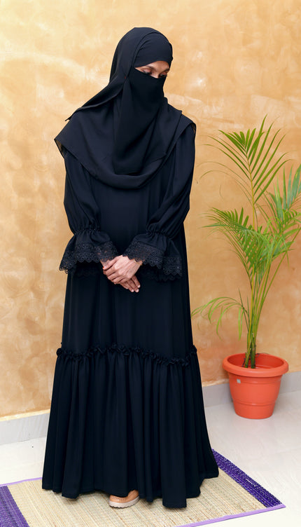 Person wearing a black abaya standing indoors with a plant in the background