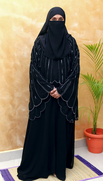 Woman wearing a black abaya with intricate designs in a room with a plant and beige wall.