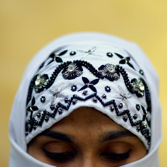 Person wearing a white hijab with black embroidery against a blurred gold background