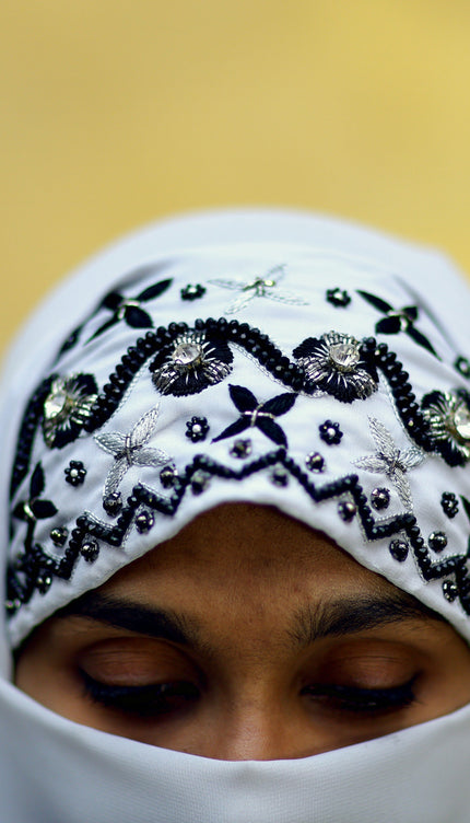 Person wearing a white hijab with black embroidery against a blurred gold background