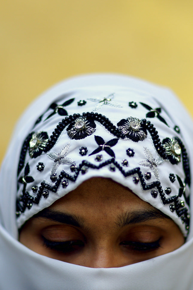 Person wearing a white hijab with black embroidery against a blurred gold background