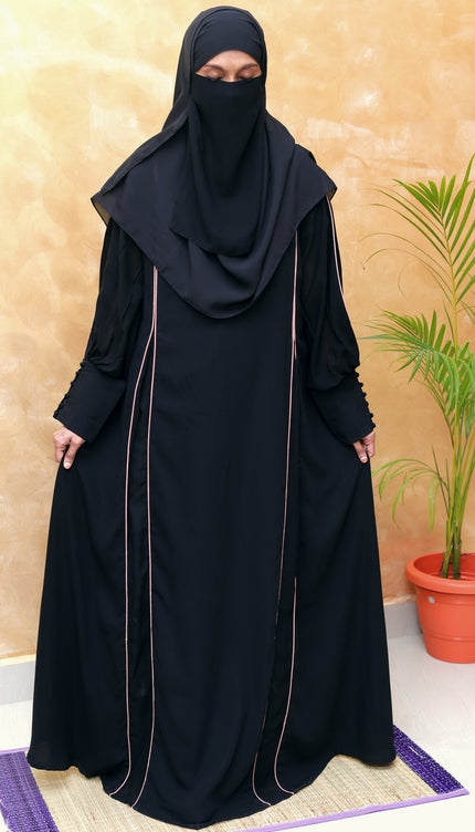 Person wearing a black abaya with a matching hijab against a beige wall.
