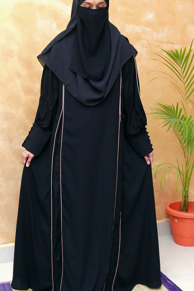 Person wearing a black abaya with a matching hijab against a beige wall.