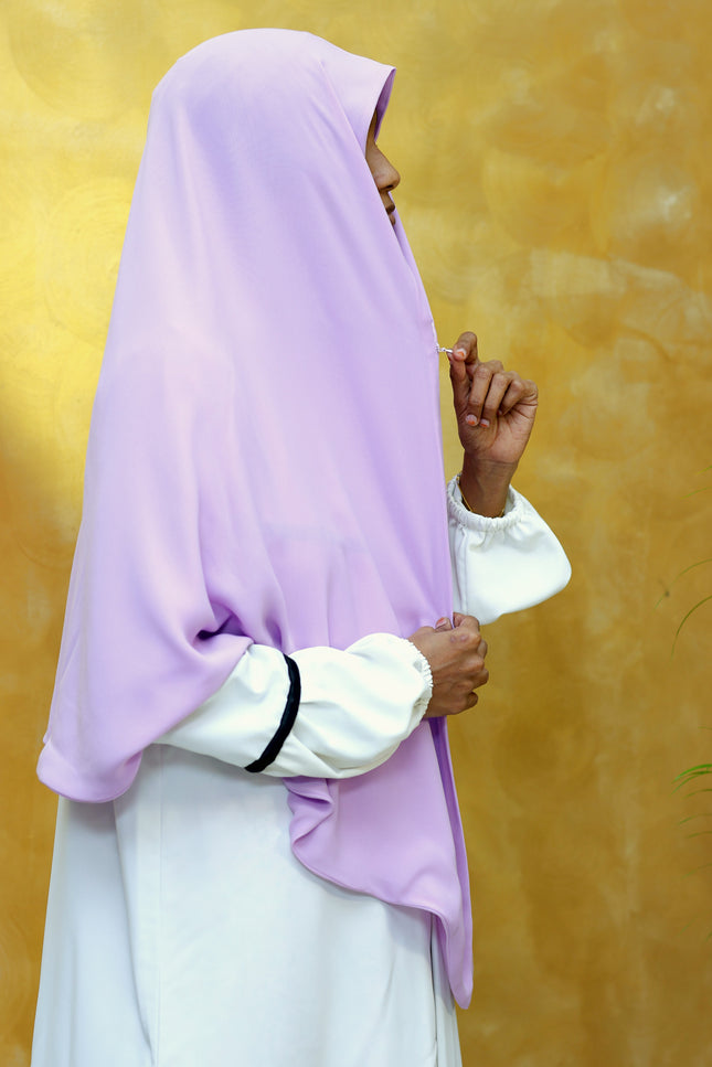 Person wearing a light purple hijab and white outfit against a yellow background