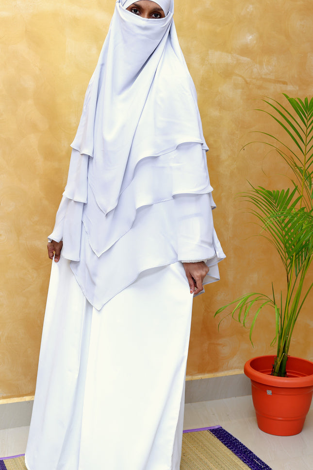 Person wearing a white layered hijab and dress against a yellow wall with a plant.