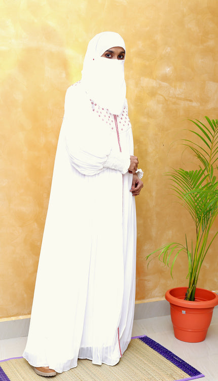 Person wearing a white abaya standing in a room with a plant and yellow wall.