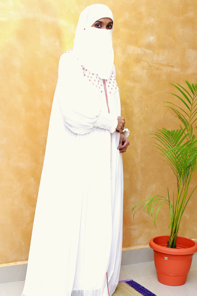 Person wearing a white abaya standing in a room with a plant and yellow wall.
